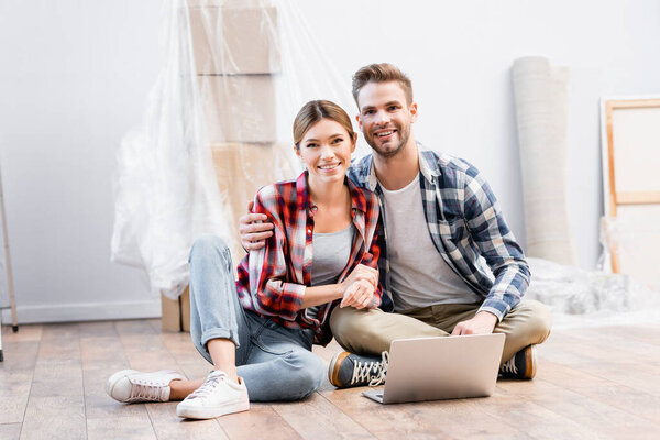 счастливая молодая пара смотрит в камеру, обнимаясь на полу рядом с ноутбуком на размытом фоне дома