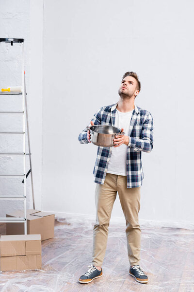 full length of young man with saucepan looking up while standing near cardboard boxes and ladder under leaking ceiling at home