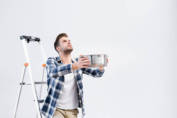 young man looking up while holding pot near ladder under leaking ceiling