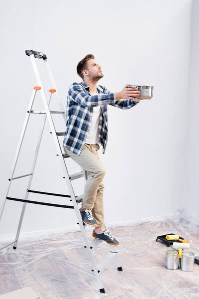 full length of young man looking up while holding pot and standing on ladder under leaking ceiling at home