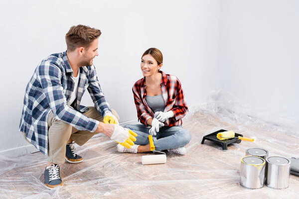 full length of happy young couple in gloves looking at each other while sitting on floor at home