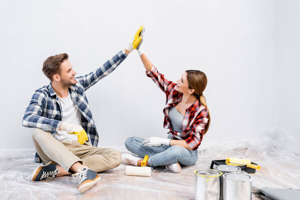 full length of happy young couple in gloves giving each other high five while sitting on floor near tins of paint at home