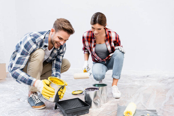full length of young man holding tin with paint while squatting near roller tray and woman at home