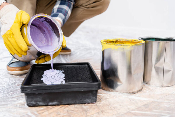 cropped view of young man pouring paint from tin in roller tray on floor indoors