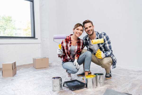 full length of happy young couple with rollers looking at camera while squatting near tins of paint and tray at home