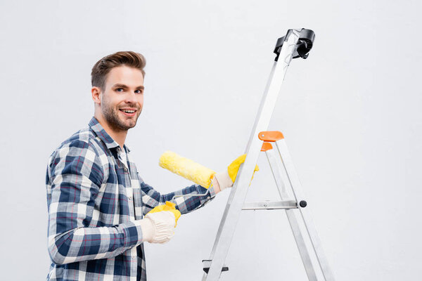 happy young man looking at camera while holding paint roller on ladder isolated on white