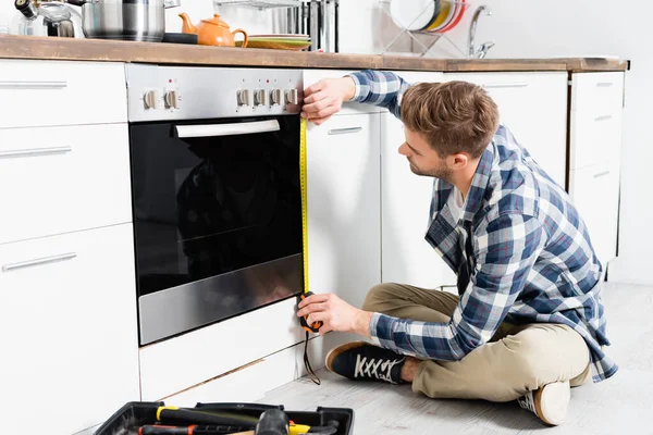 Full Length Young Man Tape Measuring Oven While Sitting Toolbox — Stock Photo, Image