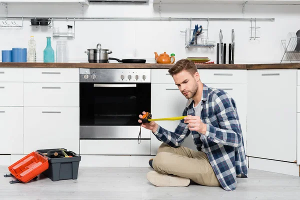 オーブンや工具箱の近くの床に座っている間にテープ測定を見ている焦点を当てた若い男の完全な長さ — ストック写真