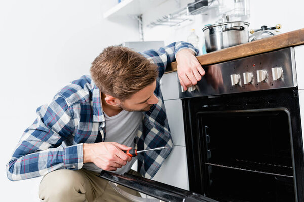 young man with screwdriver looking in oven on blurred background in kitchen