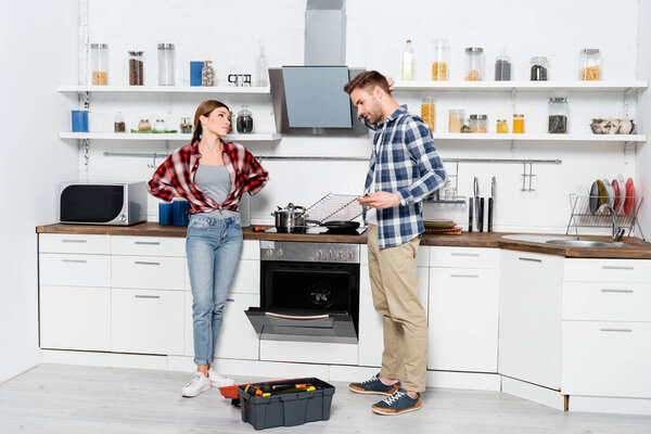 full length of dissatisfied woman looking at man holding grid near disassembled oven and toolkit in kitchen