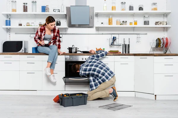 stock image full length of young woman with coffee cup sitting on table near man repairing oven in kitchen