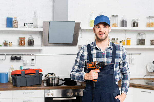 front view of happy young man with hand in pocket looking at camera while holding drill with blurred kitchen on background