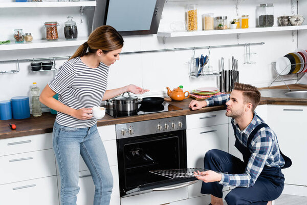 young woman with cup of coffee talking to handyman holding grid near disassembled oven in kitchen