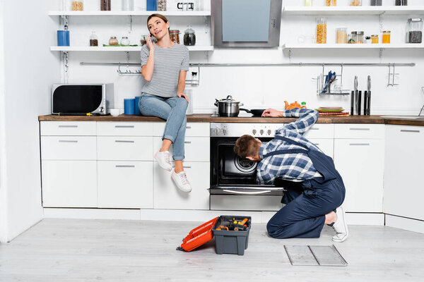full length of smiling woman talking on smartphone while sitting on table near handyman repairing oven in kitchen