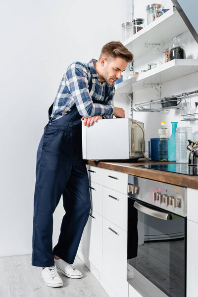 full length of young repairman looking at microwave oven in kitchen