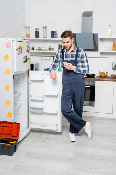 Full Length Smiling Handyman Texting Smartphone While Leaning Fridge Kitchen — Stock Photo, Image