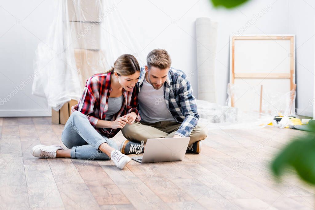 happy young couple looking at laptop while sitting on floor on blurred foreground at home