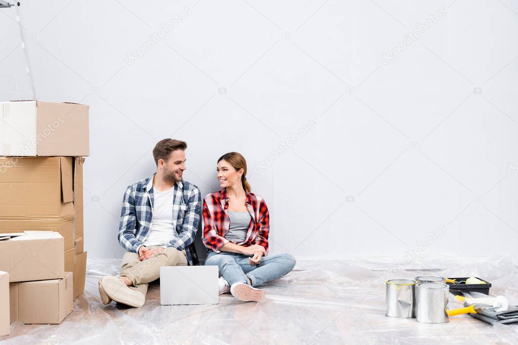 full length of happy young couple looking at each other while sitting near laptop and cardboard boxes on floor at home