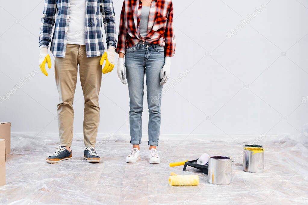 cropped view of young couple in gloves standing near tins of paint, rollers and tray on floor covered with polyethylene 