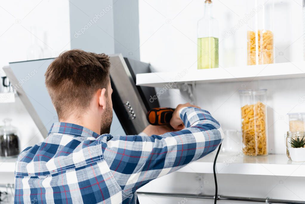 back view of young man with drill repairing extractor fan on blurred background in kitchen