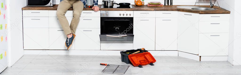 cropped view of young man sitting on table near disassembled oven and toolbox in kitchen, banner