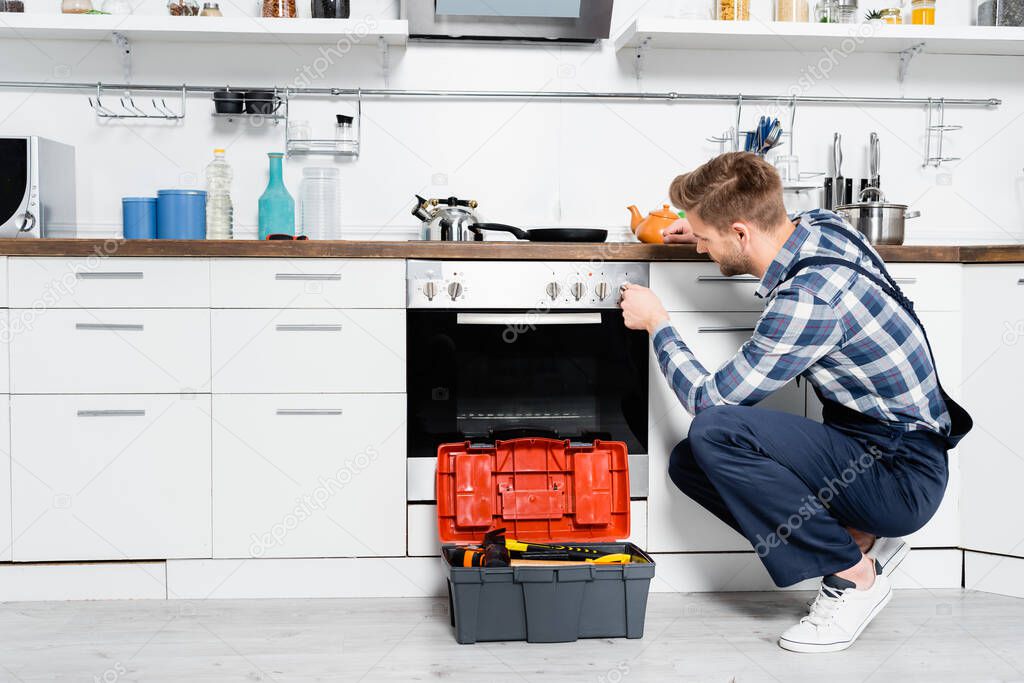 full length of young repairman turning oven button while sitting near toolbox in kitchen