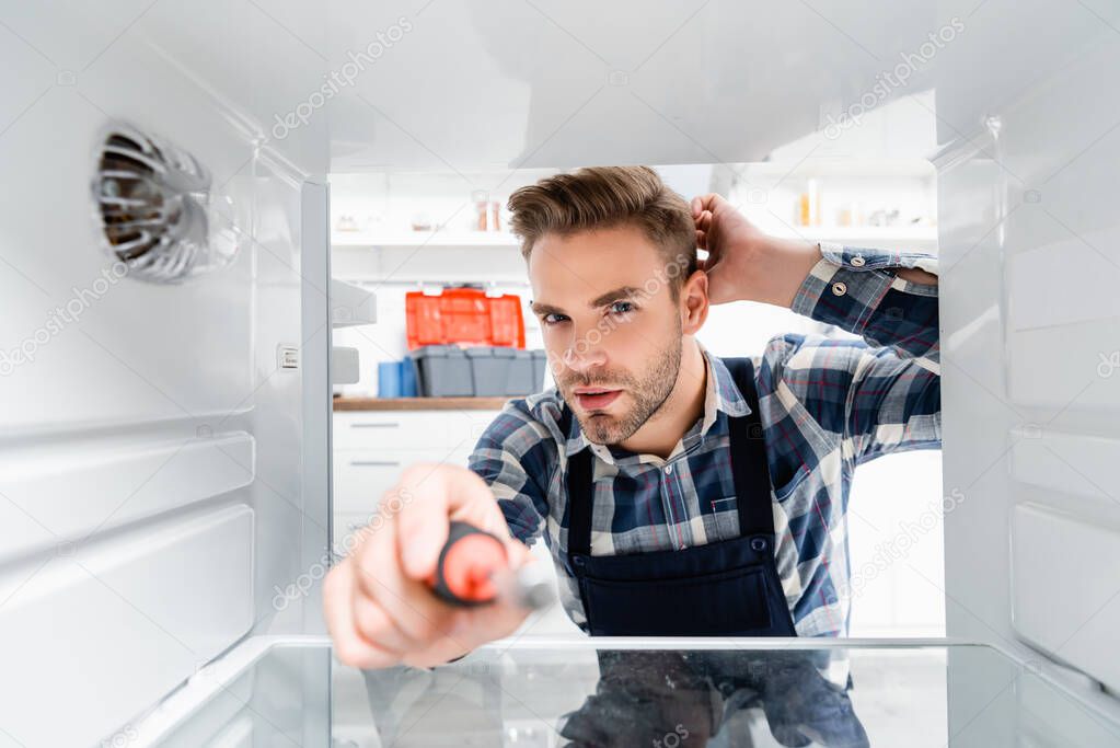 young thoughtful repairman with screwdriver looking at camera in freezer on blurred foreground