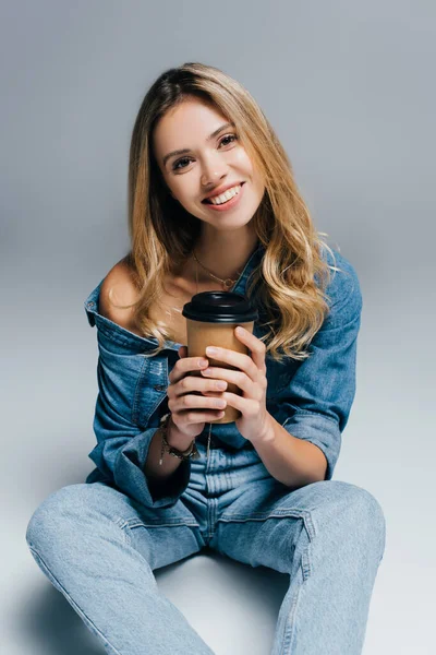 happy young woman in denim clothes, with naked shoulder, holding coffee to go while sitting on grey