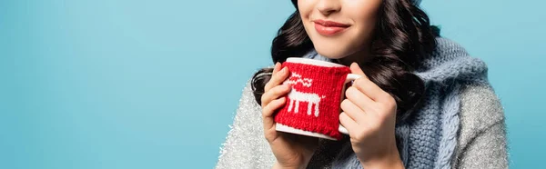 Cropped view of brunette woman in scarf holding cup with knitted holder isolated on blue, banner