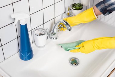 Cropped view of woman in rubber gloves wetting rag near detergent on sink in bathroom  clipart