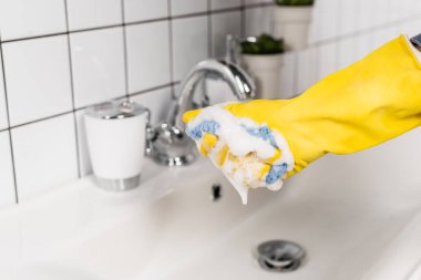 Kauçuk eldivenli, süngerli, lavabonun yanındaki bulanık arka planda banyo köpüğü tutan insan görüntüsü. 