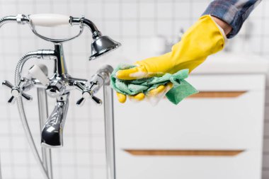Cropped view of woman in rubber glove holding rag with soap near faucet and shower in bathroom  clipart