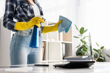 Lastik eldivenli, dizüstü bilgisayarın yanında bez ve deterjan tutan ve ön planda bulanık hesap makinesi olan kadın görüntüsü. 