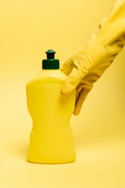 Lastik eldivenli el görüntüsü sarı arka planda bulaşık deterjanı alıyor.