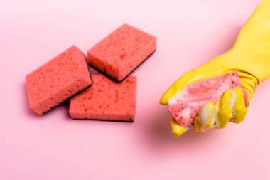 Top view of hand in rubbed glove holding sponge with soapsuds near sponges blurred on pink background clipart