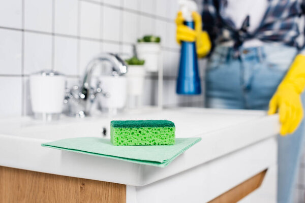Green rag and sponge on sink near woman in rubber gloves cleaning bathroom on blurred background 