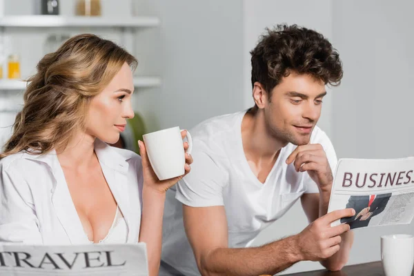 带着杯子的性感女人看着男朋友在厨房看商业报纸 — 图库照片