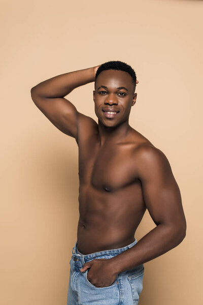 Африканский мужчина без рубашки улыбается, стоя с рукой в кармане, изолированном от бежевого