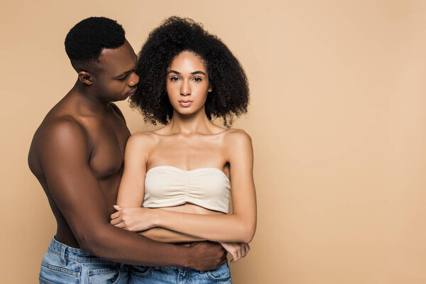 African american man hugging curly girlfriend with crossed arms isolated on beige