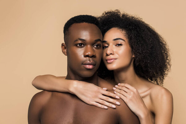 Curly african american woman with bare shoulders hugging shirtless boyfriend isolated on beige