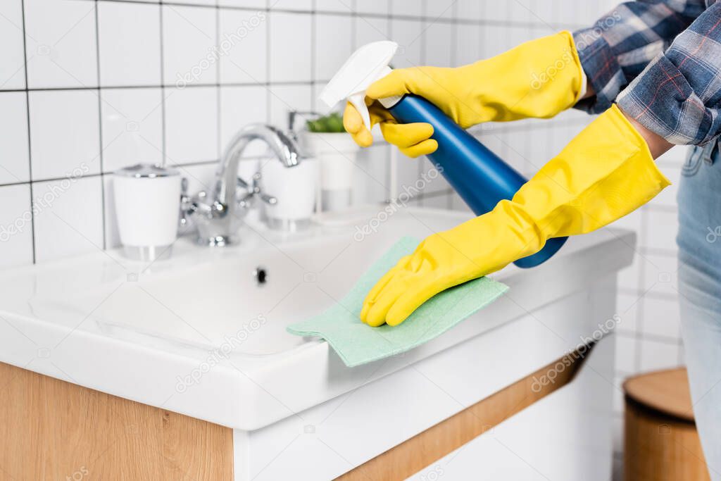 Cropped view of young woman in rubber gloves cleaning sink with detergent and rag in bathroom 
