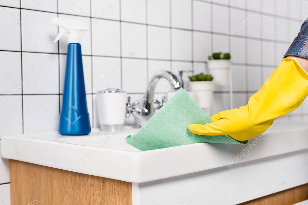 Cropped view of woman in rubber glove cleaning bathroom sink with rag near bottle of detergent on blurred background 