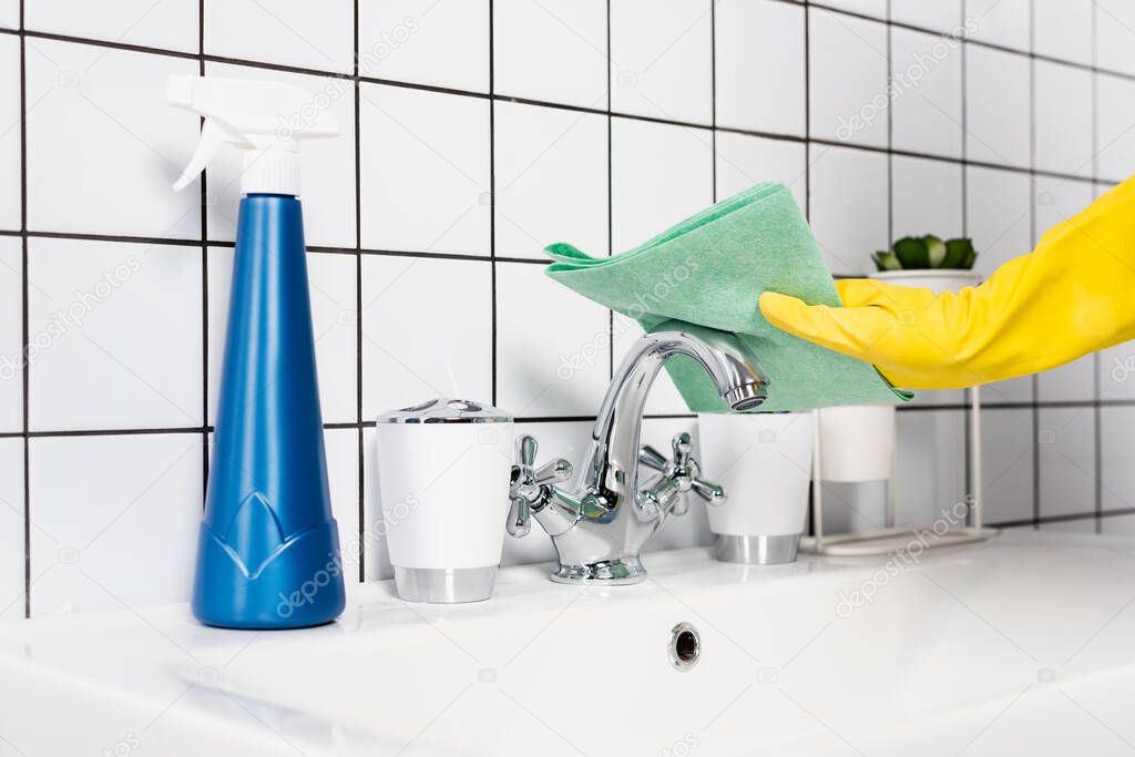 Cropped view of person in rubber glove cleaning faucet with rag near bottle of detergent on sink in bathroom 