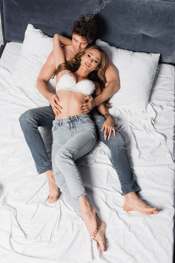 Top view of shirtless man in jeans touching seductive woman looking at camera on bed 