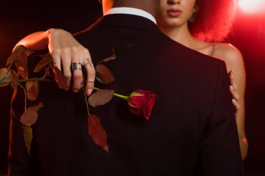 cropped view of african american woman holding rose and embracing man in formal wear on black clipart