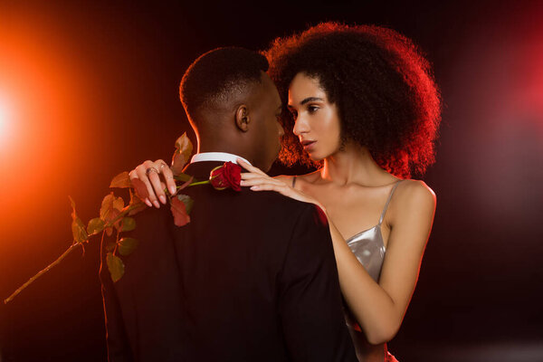 elegant african american woman holding red rose and embracing man in formal wear on black