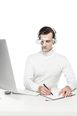 Kulaklıklı Cyborg adam ve dijital göz merceği bilgisayarın yanında, beyaz masadaki bilgisayarın üzerinde yazıyor.