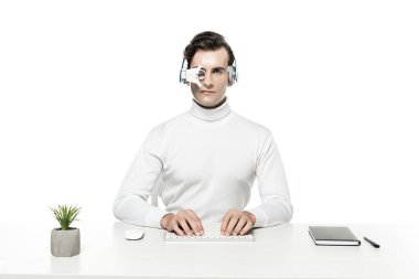 Kulaklık ve göz merceğindeki cyborg bitkinin yanında bilgisayar klavyesi ve beyaz üzerine izole edilmiş defter kullanıyor.