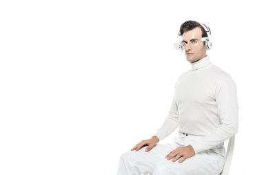 Kulaklıklı ve gözlüklü Cyborg adam beyaz sandalyede izole bir şekilde otururken kameraya bakıyor.