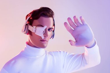Cyborg in digital eye lens and headphones looking at camera near hand on blurred foreground on purple background clipart